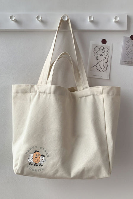 Happy Stack Cookies merch tote bag with a short and long strap, hanging off a hook.